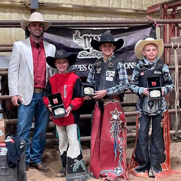 A group of boys donning cowboy hats and shirts hold up their award custom belt buckles with pride.